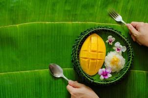 Thai dessert - Mango sticky rice that put on banana leaf plate with hand holding fork and spoon. photo