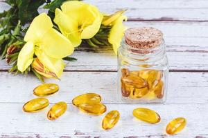 cápsulas de gel de aceite de onagra en botella de vidrio, pastillas y flores amarillas en la mesa de madera foto