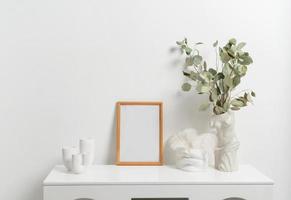 Wooden vertical frame with white vase of eucaliptus leaves over white wall photo
