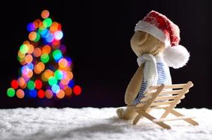 Brown teddy bear wearing scarf and santa hat sitting alone on wooden bench with snow in winter looking at colorful bokeh lights of Christmas tree. photo