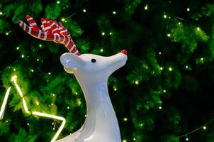 reno blanco con asta roja se para frente a la decoración del árbol de navidad con luces. foto