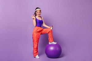 Pretty girl in great mood put her foot on fitball and poses with bar of chocolate on purple backgro