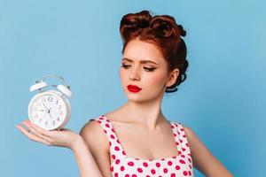 Sad woman with pinup hairstyle looking at clock. Charming sad girl in polka-dot dress posing on blu