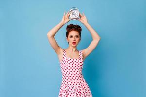 Unhappy young woman holding clock. Studio shot of upset ginger girl in polka-dot dress.