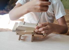 el primer plano de la mano de un niño monta un coche de juguete de madera con un destornillador en una mesa de madera en casa. foto