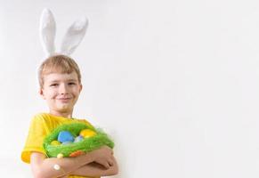 niño feliz sonriendo en una camiseta amarilla en orejas de conejo y con huevos coloridos en una canasta sobre un fondo blanco. feliz concepto de pascua con lugar para el texto. foto