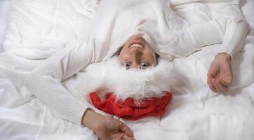 una hermosa morena yace en una cama blanca con un sombrero de santa claus mira la cámara y sonríe. Feliz Navidad. foto