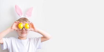 A happy child in a white t-shirt in bunny ears and with colorful yellow Easter eggs in front of his eyes on a white background. Happy Easter concept. photo
