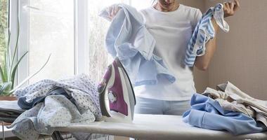 Frustrated tired young woman holds stares at iron and pile of ironed clothes at home. Housework photo