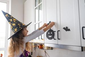 niño decora la cocina en casa para halloween. una chica disfrazada de bruja juega con la decoración de las vacaciones: murciélagos, farolillos, calabazas. comodidad otoñal en casa, cocina de estilo escandinavo, loft foto