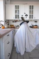 A child in sheets with cutout for eyes like a ghost costume in the kitchen decorated for the Halloween holiday. A kind little funny ghost. Halloween Party photo