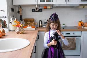 Child decorates the kitchen in home for Halloween. Girl in a witch costume plays with the decor for the holiday - bats, jack lantern, pumpkins. Autumn comfort in house, Scandi-style kitchen, loft photo