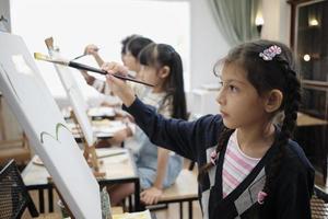 una niña pequeña se concentra en la pintura acrílica en color sobre lienzo con niños multirraciales en un aula de arte, aprendizaje creativo con talentos y habilidades en la educación de estudio de la escuela primaria. foto