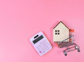 flat layout of wooden house model in shopping trolley and pink calculator on pink  background with copy space, home purchase concept. photo