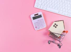 flat layout of wooden house model in shopping trolley and pink calculator and computer keyboard  on pink  background with copy space, home purchase concept. photo