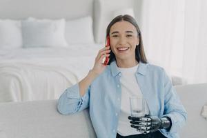 Smiling disabled girl speak by phone using bionic arm prosthesis, holding smartphone, glass of water photo