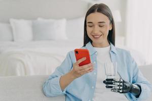 Disabled girl with bionic prosthetic arm using mobile phone apps at home, holding glass of water photo