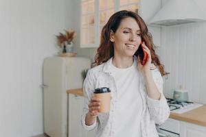 Smiling female holding takeaway coffee, talking on phone, enjoying pleasant conversation in kitchen