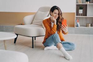 Modern girl holding smartphone taking selfie photo or chatting by video call sitting on heated floor