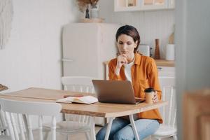 Pensive female freelance worker businesswoman works online at laptop in kitchen at home. Remote job photo