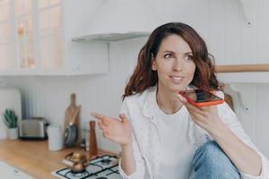 Happy hispanic female sends audio voice message, chatting, using speaker phone in kitchen at home photo