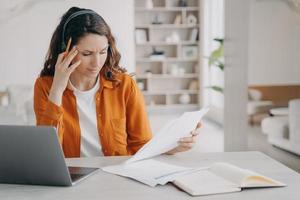 Female employee in headset looking through paper document, working at laptop at home. Remote work photo