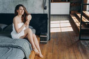 Girl wrapped in towel combs long hair after morning shower sitting on bed. Natural beauty, haircare photo