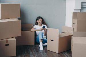 Happy girl choosing moving company for removal using phone, sitting with boxes. Mortgage, relocation photo