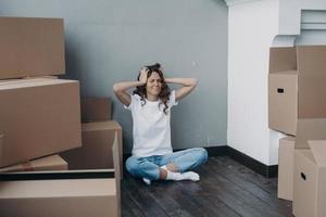Tired stressed girl holding her head sitting with boxes having problem with relocation on moving day photo