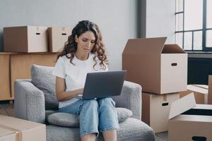 chica comprando en línea una nueva casa en una computadora portátil, sentada entre cajas de tarjetas. reubicación, comercio electrónico foto