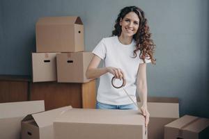 Smiling spanish woman packing stuff in cardboard box, using adhesive tape. Delivery, removal service