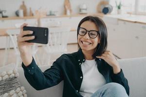 Smiling girl blogger holding smartphone, talks by video call, takes selfie photos, sitting on couch