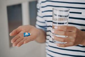 Hand of woman is holding white and blue capsules and glass of water. Lady is going to take drugs. photo