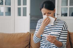Unhealthy female blowing her nose holds thermometer, suffering from high temperature, fever at home