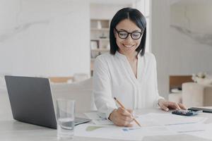 una mujer de negocios sonriente con anteojos trabaja calculando el presupuesto del proyecto sentada en el escritorio de la oficina con una laptop foto