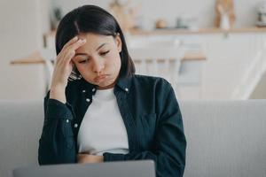 mujer independiente cansada y con exceso de trabajo que trabaja en una computadora portátil, sosteniendo la cabeza lee el correo electrónico con malas noticias en casa foto