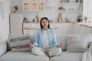 Serene woman practices yoga meditation sitting on cozy couch at home. Wellness, stress relief