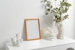 Wooden vertical frame with white vase of eucaliptus leaves over white wall photo