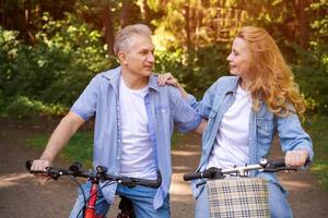 Cheerful active senior couple with bicycle walking through park together.
