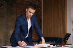 Young man in jacket working from home doing paperwork using laptop and holding photo