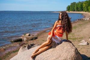 hermosa joven esbelta con cabello largo ondulado se sienta en la orilla del mar en grande foto