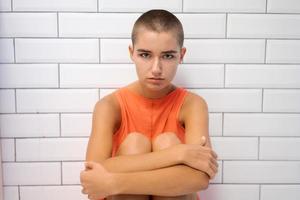 Sickly bald girl in an orange T-shirt sits on floor hugging her knees with her photo