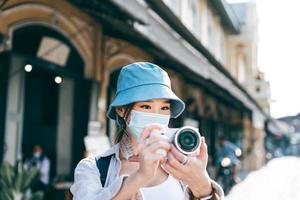 Una joven adulta asiática que viaja con mochila usa mascarilla para tomar una foto con la cámara.