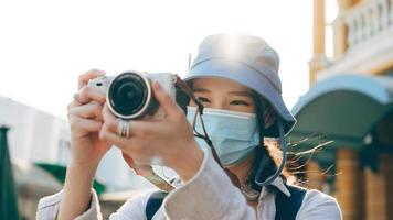 Una joven adulta asiática que viaja con mochila usa mascarilla para tomar una foto con la cámara.