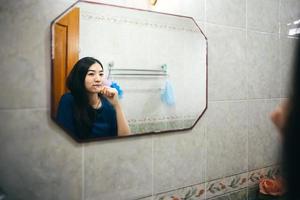 retrato de una sonrisa feliz joven adulta asiática cepillando los dientes frente al espejo en el baño foto