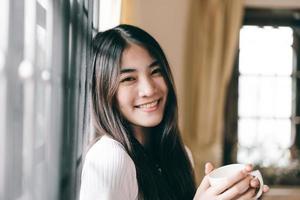 retrato de una feliz sonrisa joven adulta asiática sosteniendo una taza de café cerca de la ventana por la mañana foto