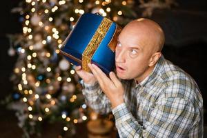 bald man listens to a box with a gift on the background of a Christmas tree photo