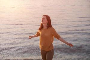 Happy woman relaxes with her arms out to sides on shore lake at sunset in rays photo