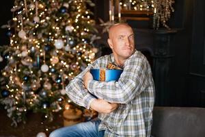 Happy bald man holding a box with a gift on the background of a christmas tree photo