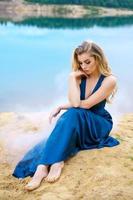 sad young beautiful woman in a blue long dress is sitting alone on the lake shore photo